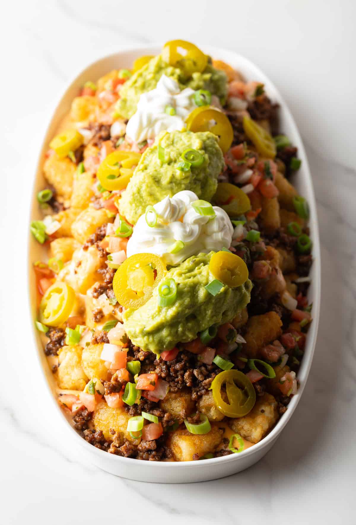 Top down view of large white oval serving dish loaded with tater tot nachos with ground beef, jalapenos, salsa, cheddar cheese, guacamole and sour cream.