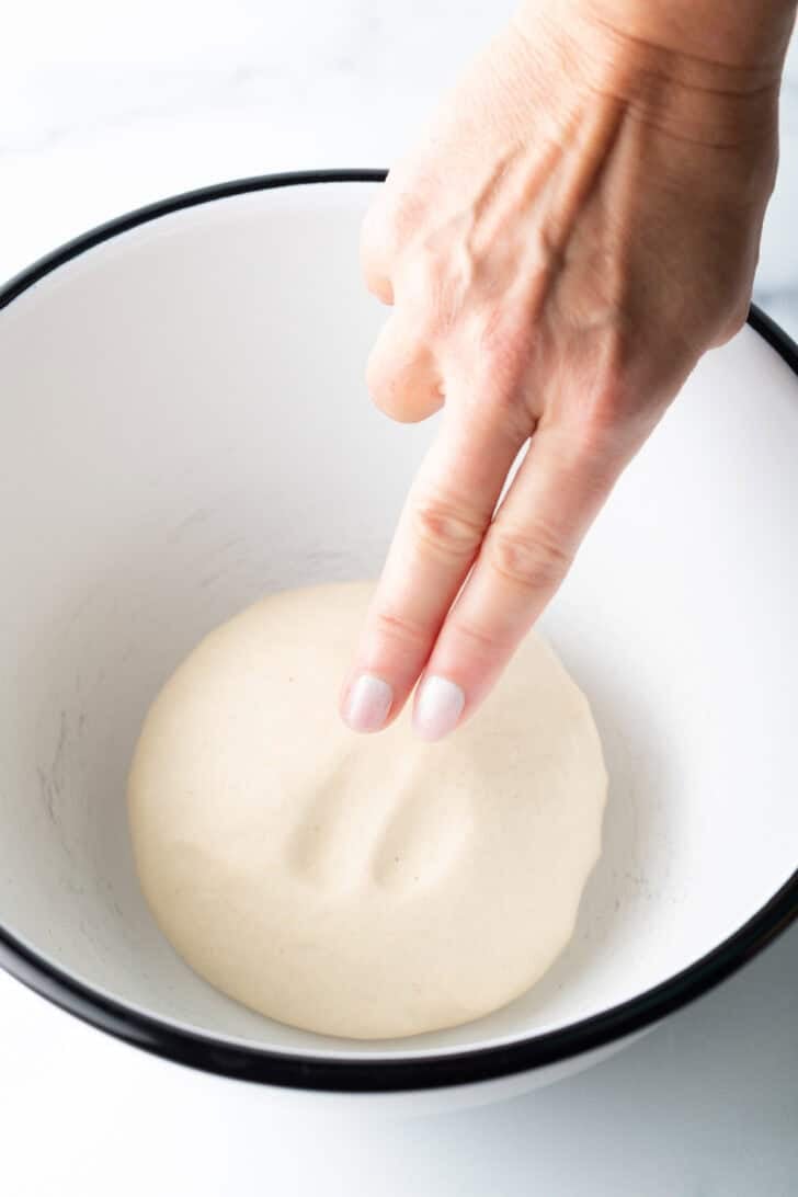 Hand using two fingers to poke a round of pizza dough in a large white mixing bowl.