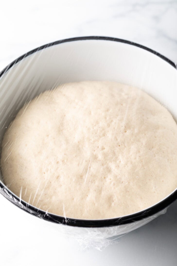 White bowl with a circle of dough covered with plastic wrap.
