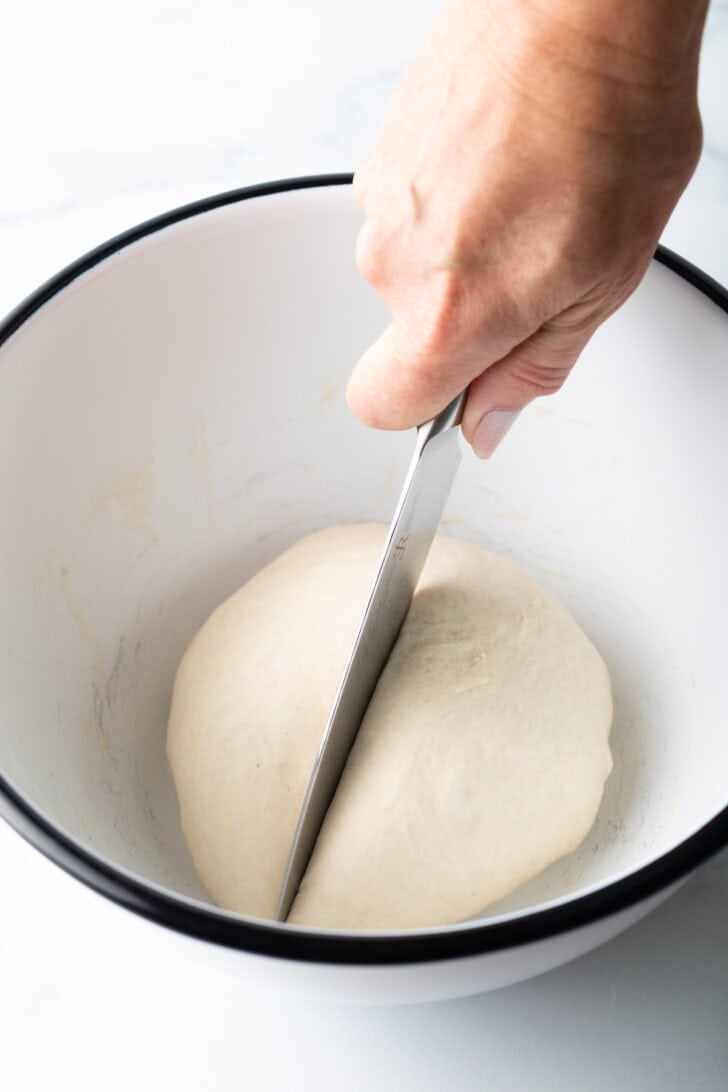 White bowl with a circle of dough. A hand is using a knife to cut the dough in half.