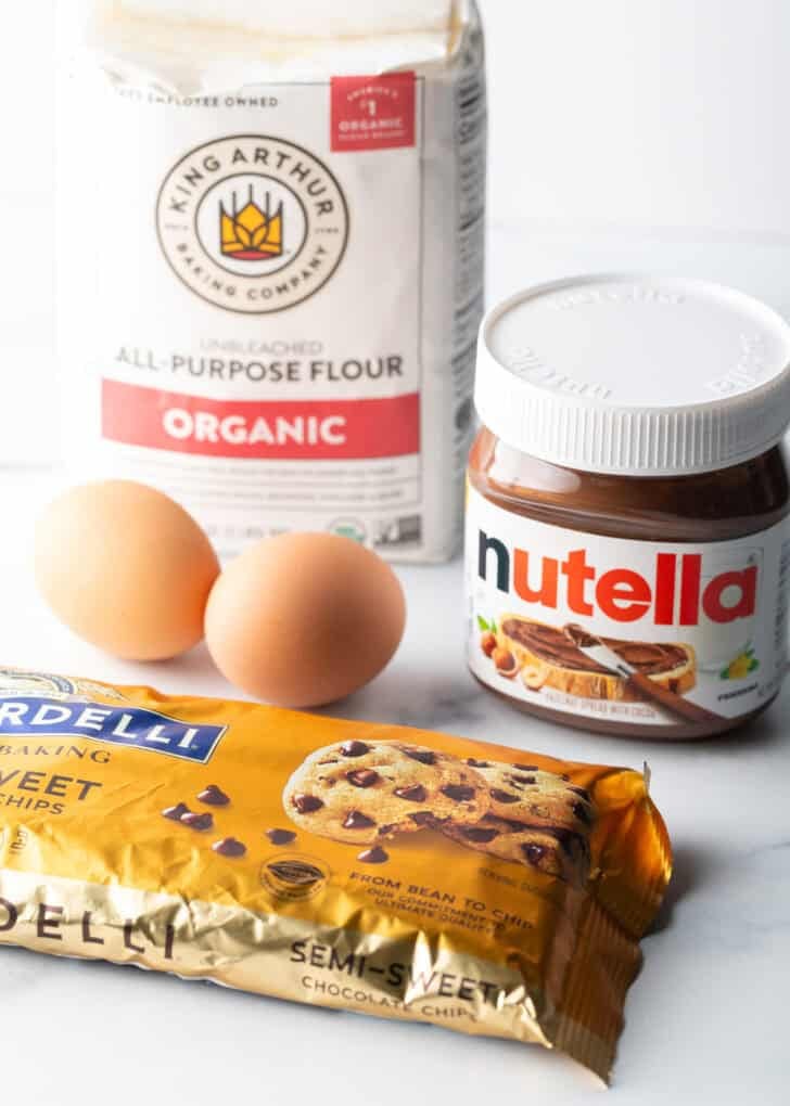 Nutella cookie recipe ingredients: Bag of chocolate chips, jar of nutella, bag of flour, and two eggs.