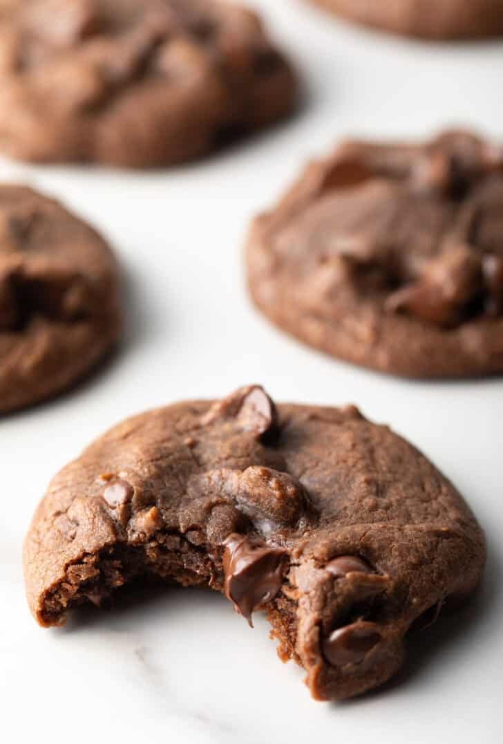 5 baked nutella cookies with chocolate chips on a white background. The closest cookie has a bite taken out of it.