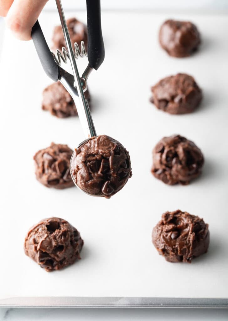 Hand with a cookie scoop showing chocolate dough to camera.