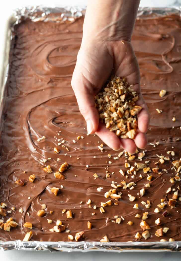 Hand sprinkling broken pieces of toffee overtop of chocolate covered saltine crackers in a baking sheet.