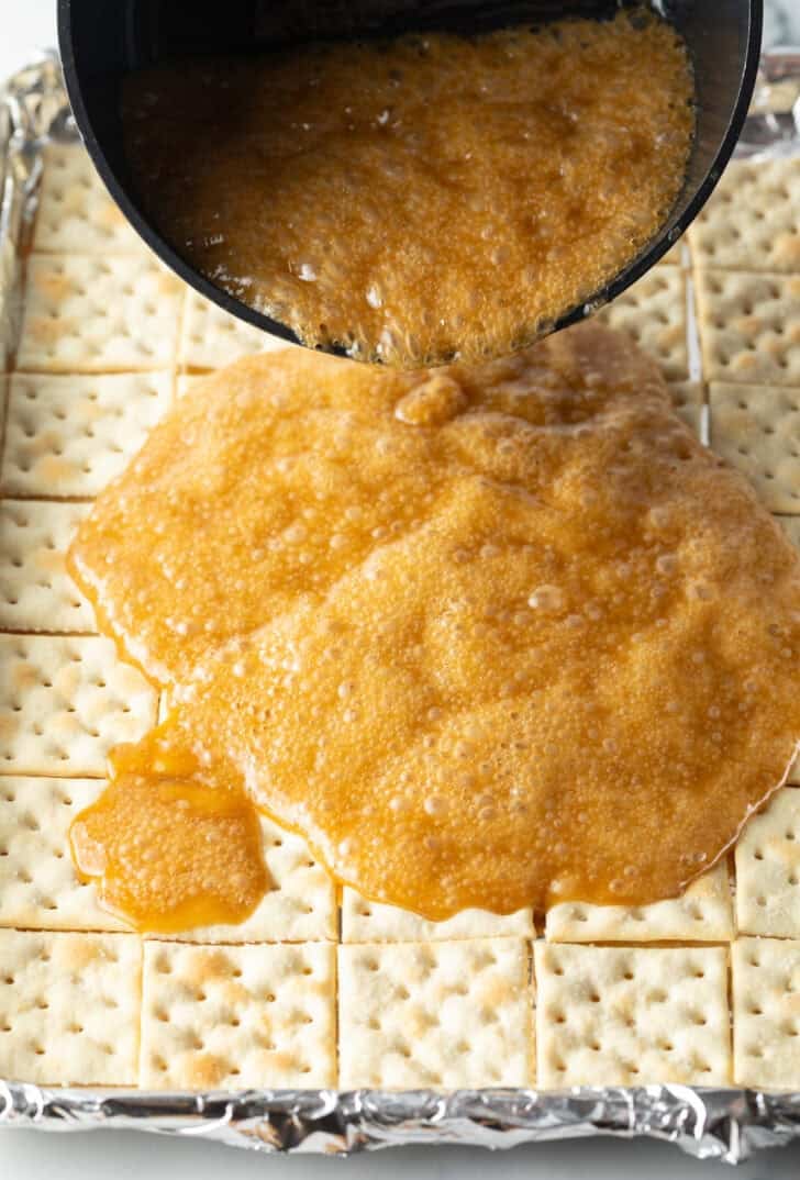 Pouring melted toffee over saltine crackers spread evenly in a baking sheet.