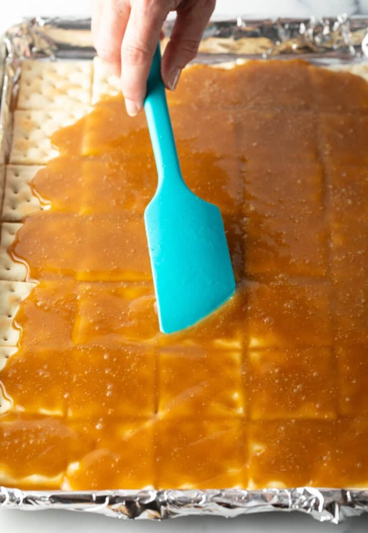 Using a blue spatula to smooth melted toffee across saltine crackers on a baking plan.