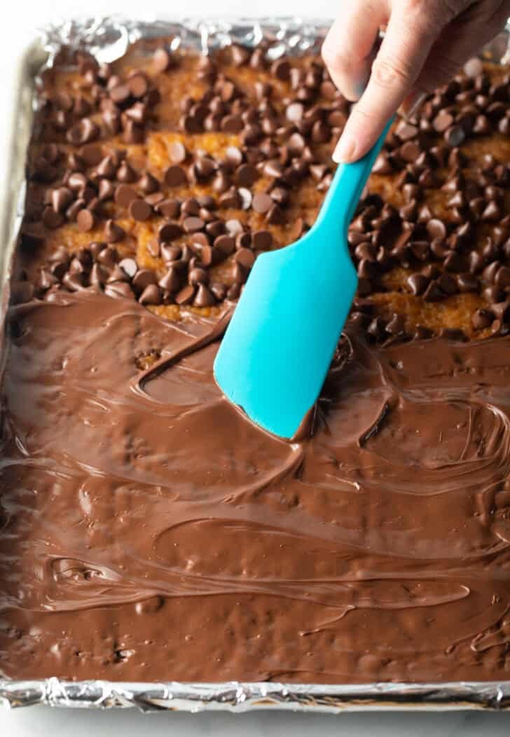Using a blue spatula to spread melted chocolate over saltine crackers in a baking sheet.