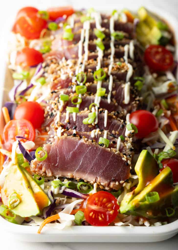 Large white rectangle dish loaded with salad greens, halved cherry tomatoes, avocado slices, slices of seared ahi tuna, chopped green onions and a drizzle of dressing.