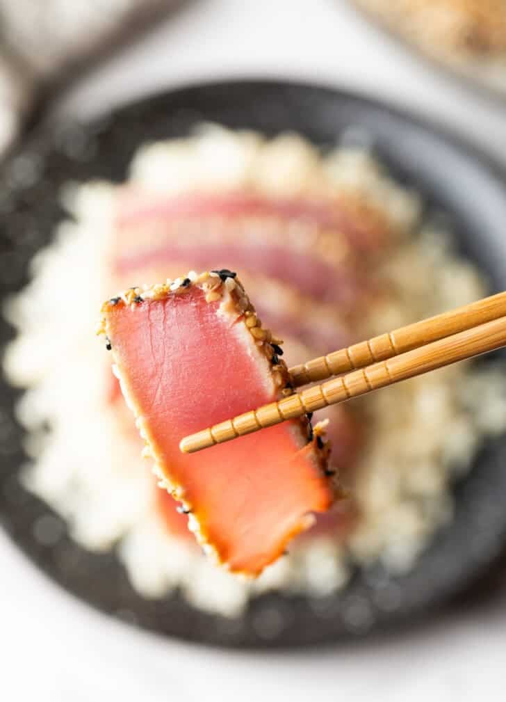 Piece of seared yellowfin tuna held with chopsticks to camera.