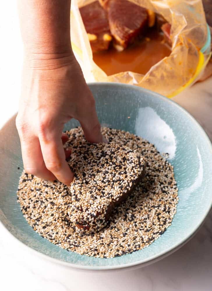 Hand pressing a raw piece of ahi tuna steak into a bowlful of tricolor sesame seeds.