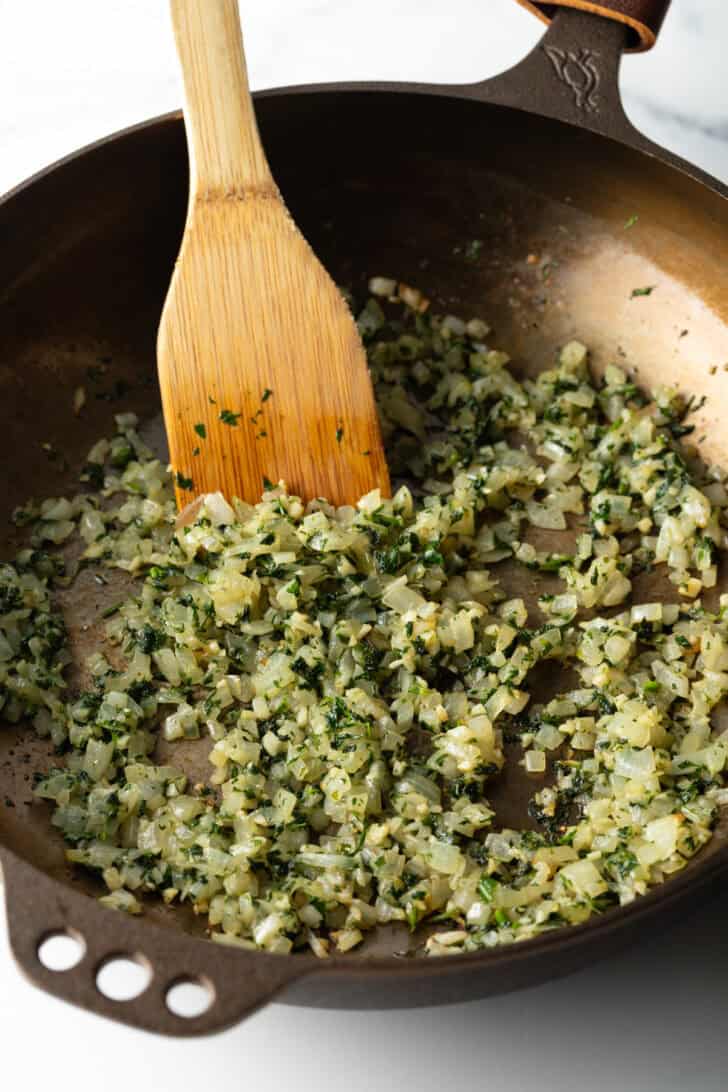Using a wooden spatula to cook onions and seasonings in a large cast iron pan.
