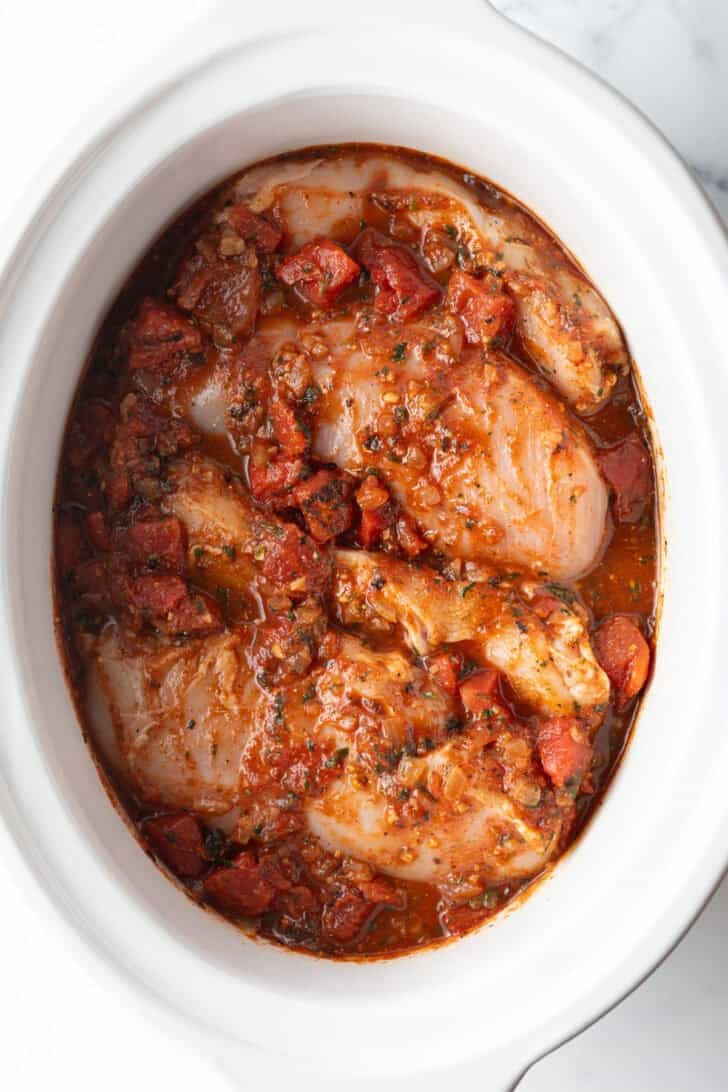 Several whole chicken breasts covered in red sauce in a white crockpot.
