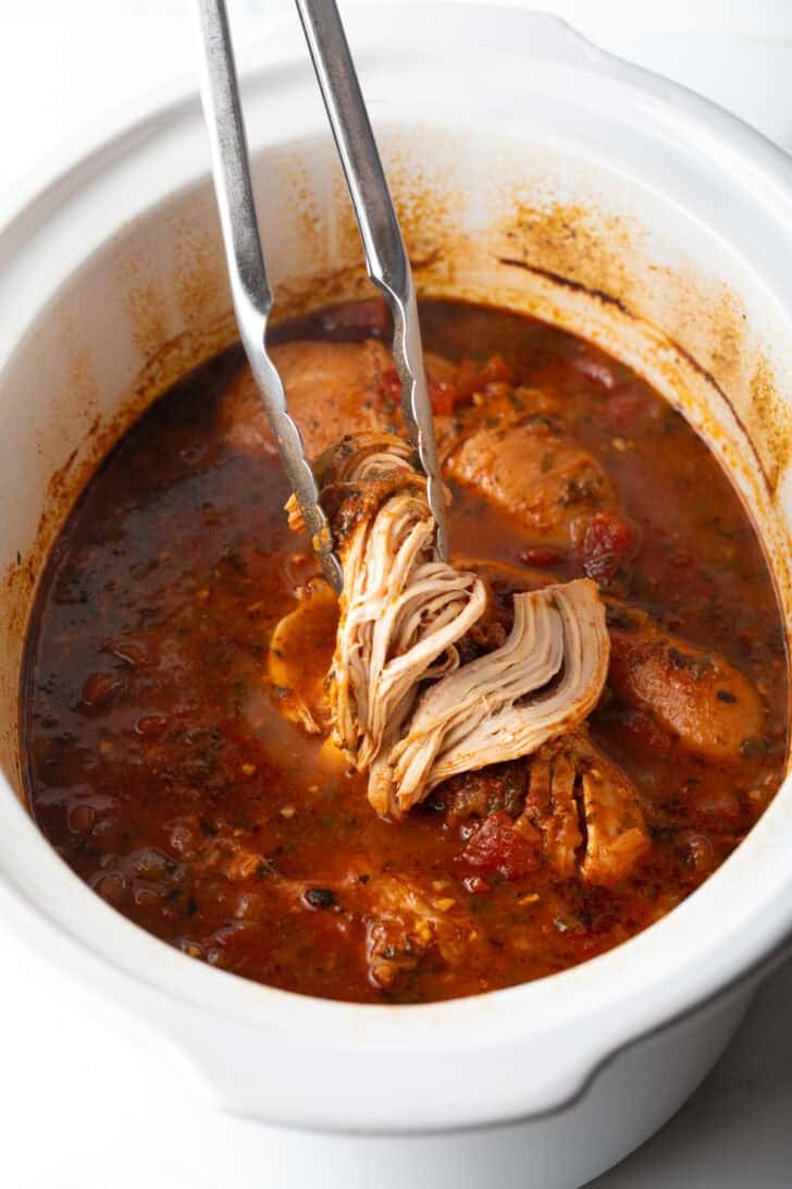 Tongs holding up shredded chicken above a white slow cooker.