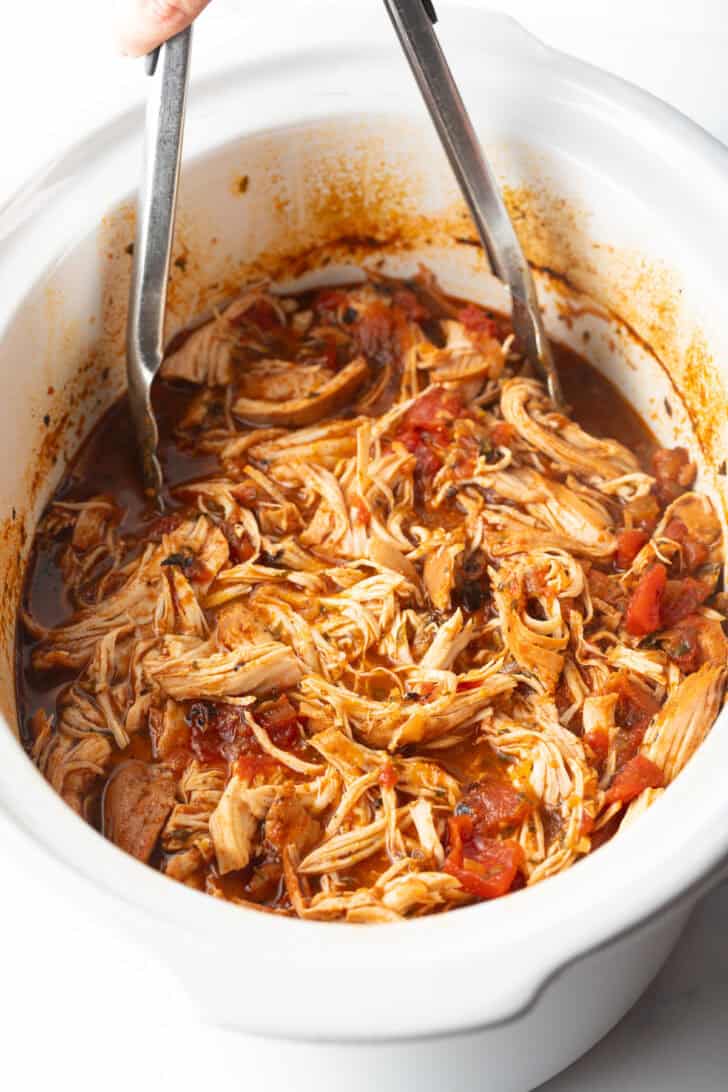 Shredded Mexican chicken in a white crockpot with tongs.