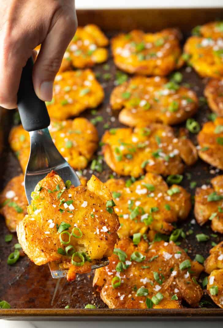 Top down view of 15 golden smashed potatoes on a baking sheet. A hand with a spatula is holding a crispy smashed potato to camera.