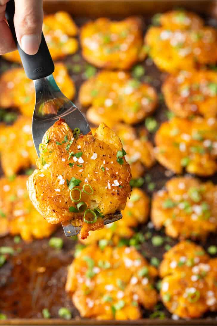 Top down view of 15 golden smashed potatoes on a baking sheet. A hand with a spatula is holding a crispy smashed potato to camera.