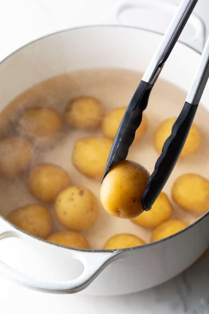Tongs holding a small boiled potato over a large white pot with a dozen small potatoes.