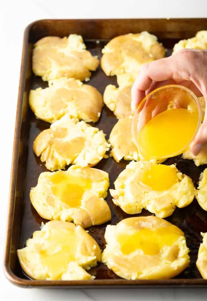 Hand pouring melted ghee from a small glass bowl over unbaked smashed potatoes on a baking sheet.