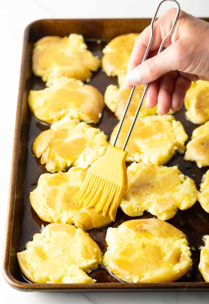 Hand using a yellow basting brush to brush ghee over smashed potatoes on a baking sheet.