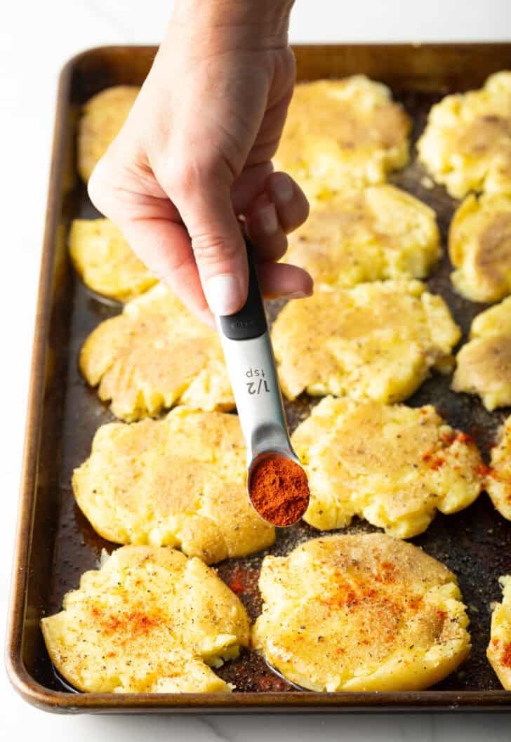 Hand adding spices from a small measuring spoon over smashed potatoes on a baking sheet.