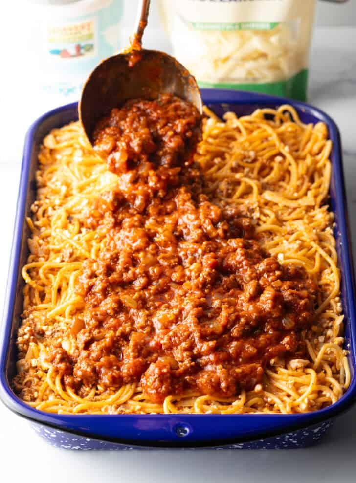 A ladle pouring meat sauce over spaghetti in a blue casserole dish.