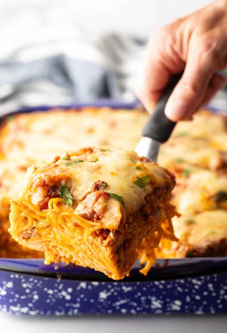 Hand with a spatula holding a serving of baked spaghetti to camera.