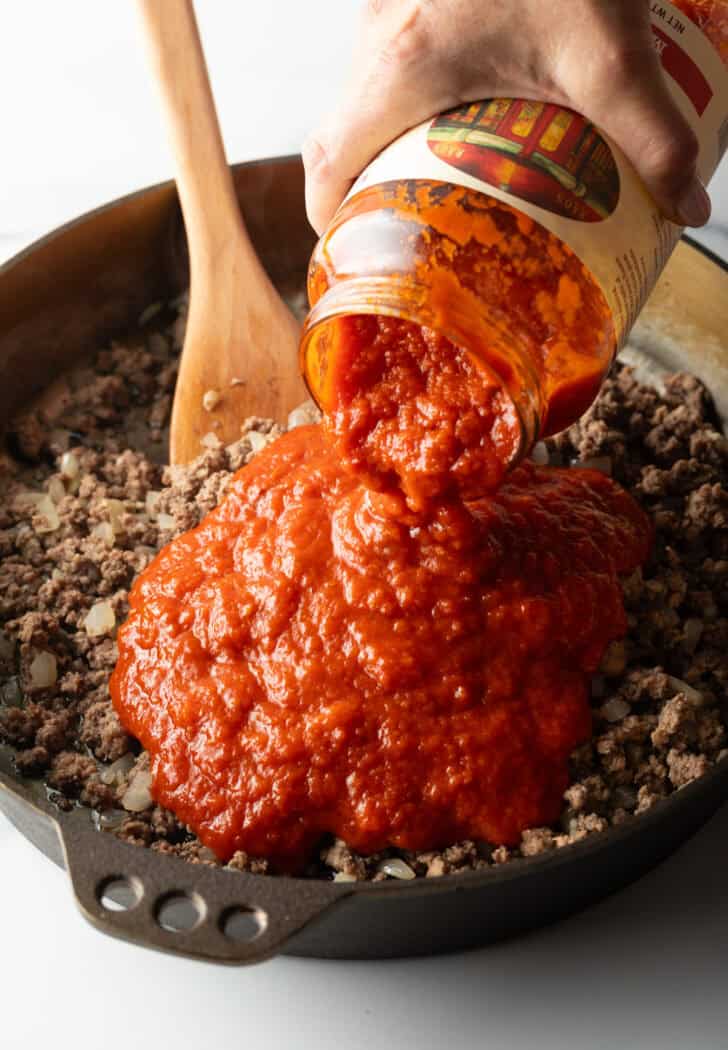 Adding jarred marinara to a skillet with ground cooked meat.