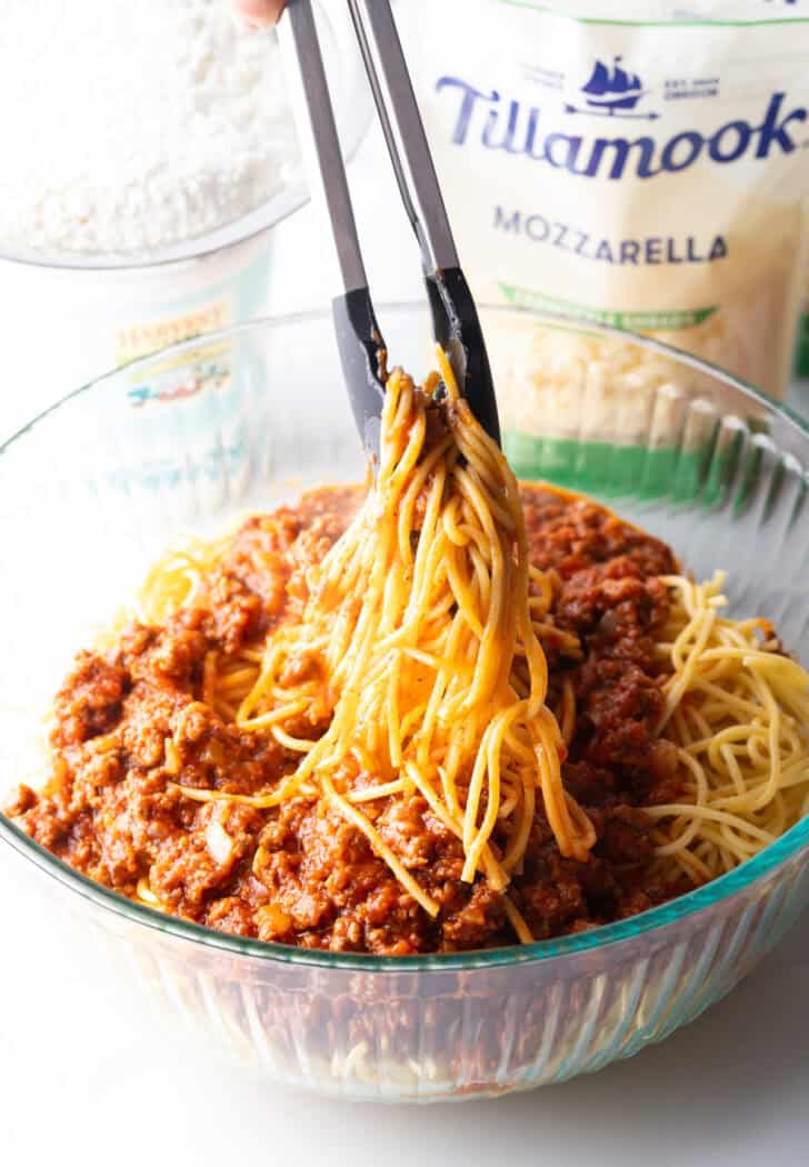 Tongs tossing spaghetti together with meat sauce in a glass bowl.