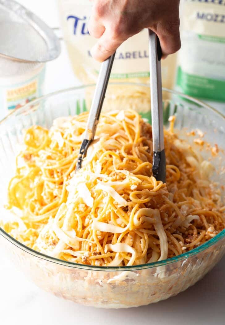 Using tongs to toss cooked spaghetti in a glass bowl.