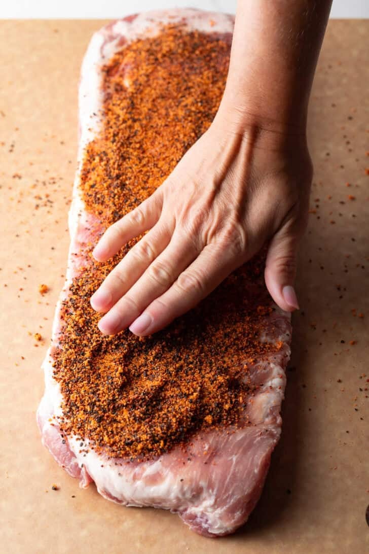 Overhead shot of a raw rack of pork ribs with a hand covering it with dry rub seasoning.