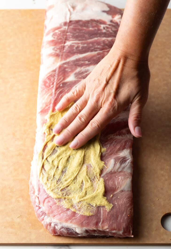 Overhead shot of a raw rack of St. Louis pork ribs being smothered with Dijon mustard.