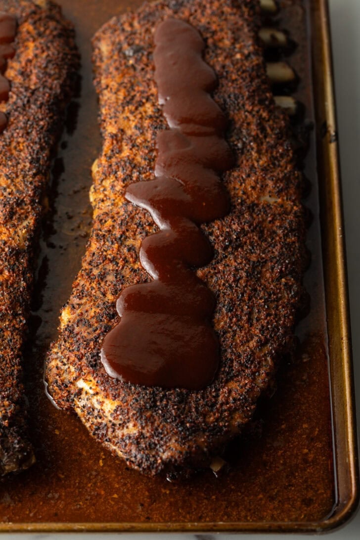 Cooked ribs on oven tray drizzled with barbecue sauce.