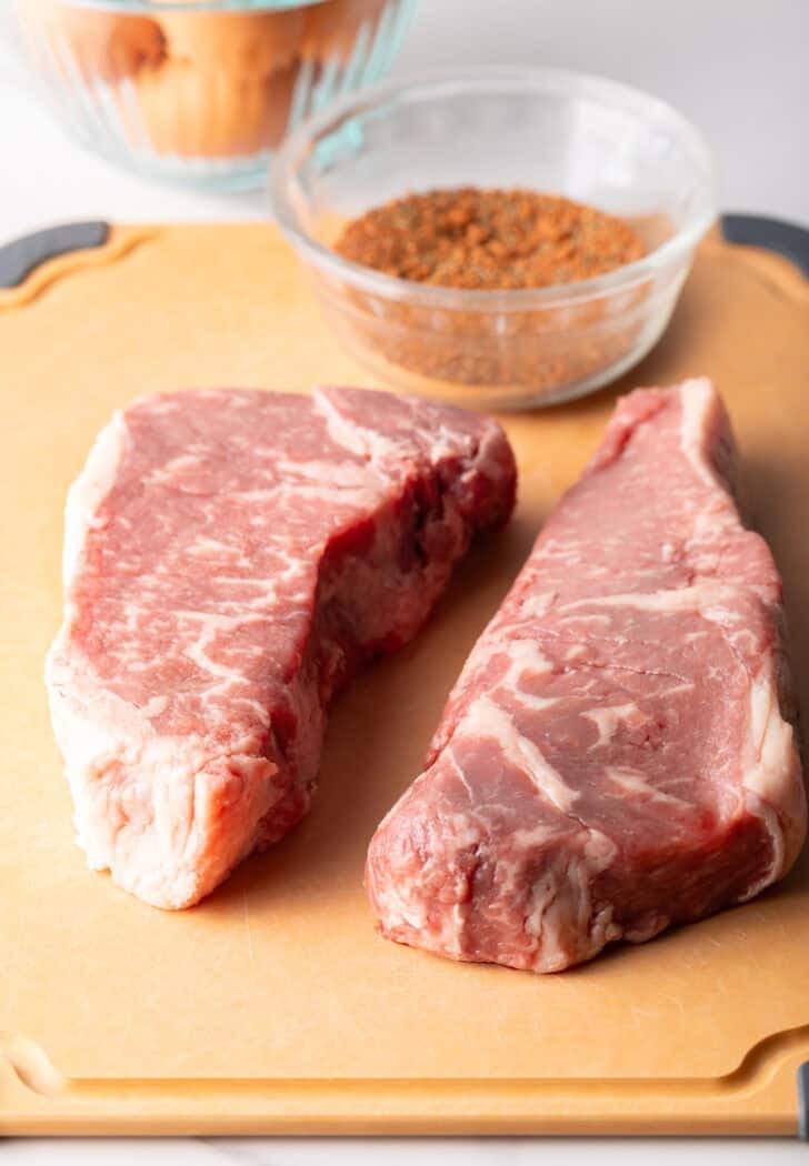 Two raw steaks and a glass bowl with seasoning blend.