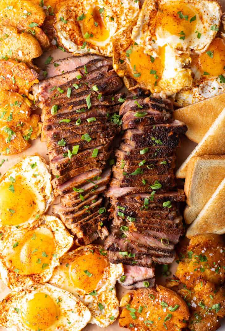 Top down view of two seared steaks side-by-side, each cut widthwise into thin strips, and surrounded by mounds of fried eggs, triangles of toast, and smashed potatoes.