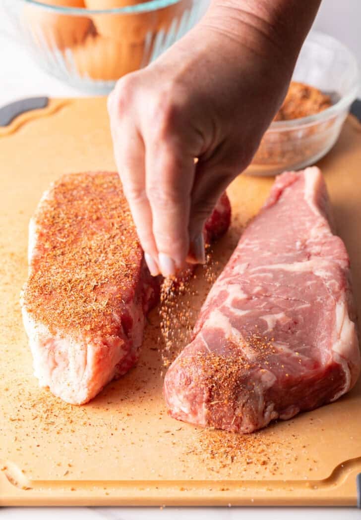 A hand sprinkling steak seasoning over two raw ribeyes.