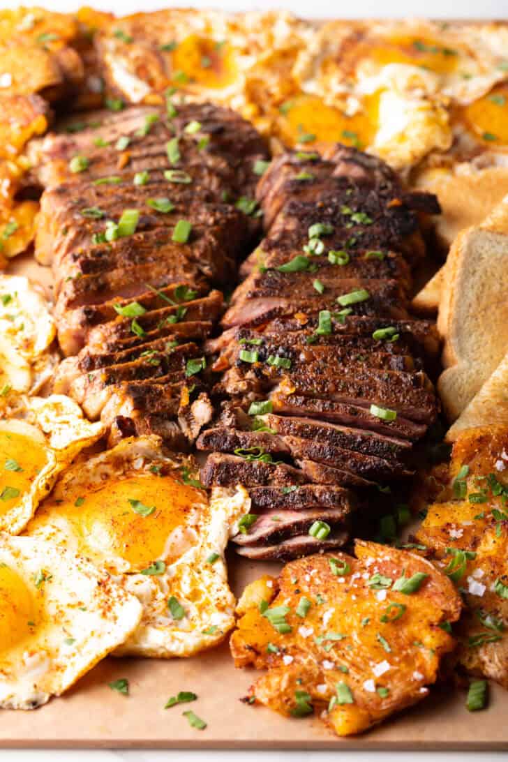 Top down view of two seared steaks side-by-side, each cut widthwise into thin strips, and surrounded by mounds of fried eggs, triangles of toast, and smashed potatoes.