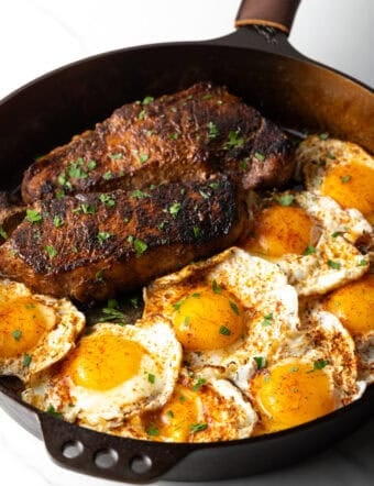 Cast iron skillet with two seared steaks and 8 fried eggs.