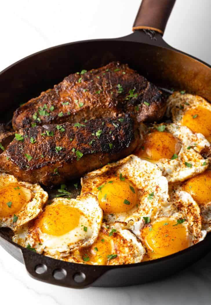 Cast iron skillet with two seared steaks and 8 fried eggs.