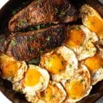 Cast iron skillet with two seared steaks and 8 fried eggs.