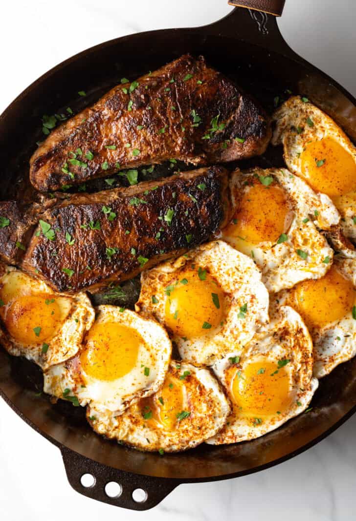 Cast iron skillet with two seared steaks and 8 fried eggs.