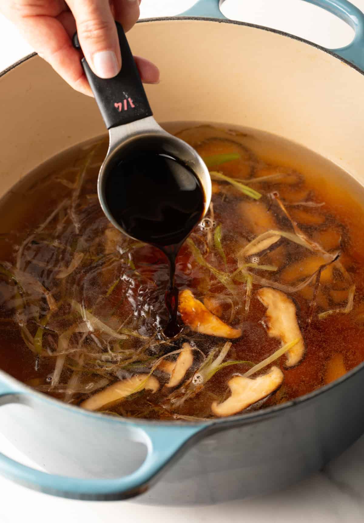 A measuring cup adding soy sauce to udon noodle soup broth.