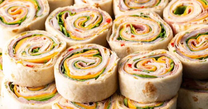 Horizontal image of a dozen stacked ham and cheese pinwheel sandwiches.