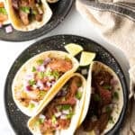 Top down view of a black plate with 3 tacos, featuring marinated steak and toppings.