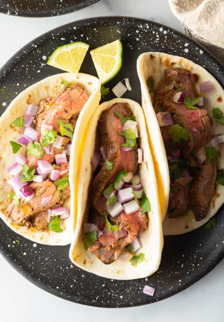 3 steak tacos on a black plate.