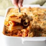 A serving of easy chicken parmesan casserole is being scooped out of a white baking dish.