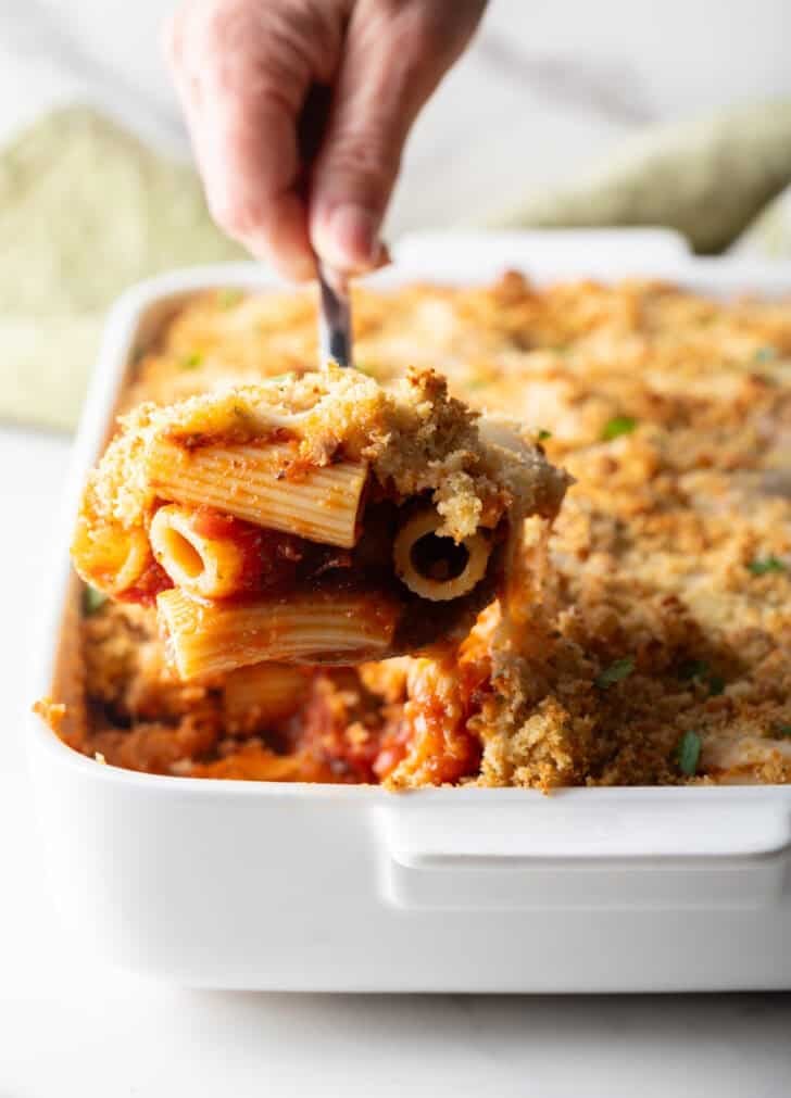 A serving of easy chicken parmesan casserole is being scooped out of a white baking dish.