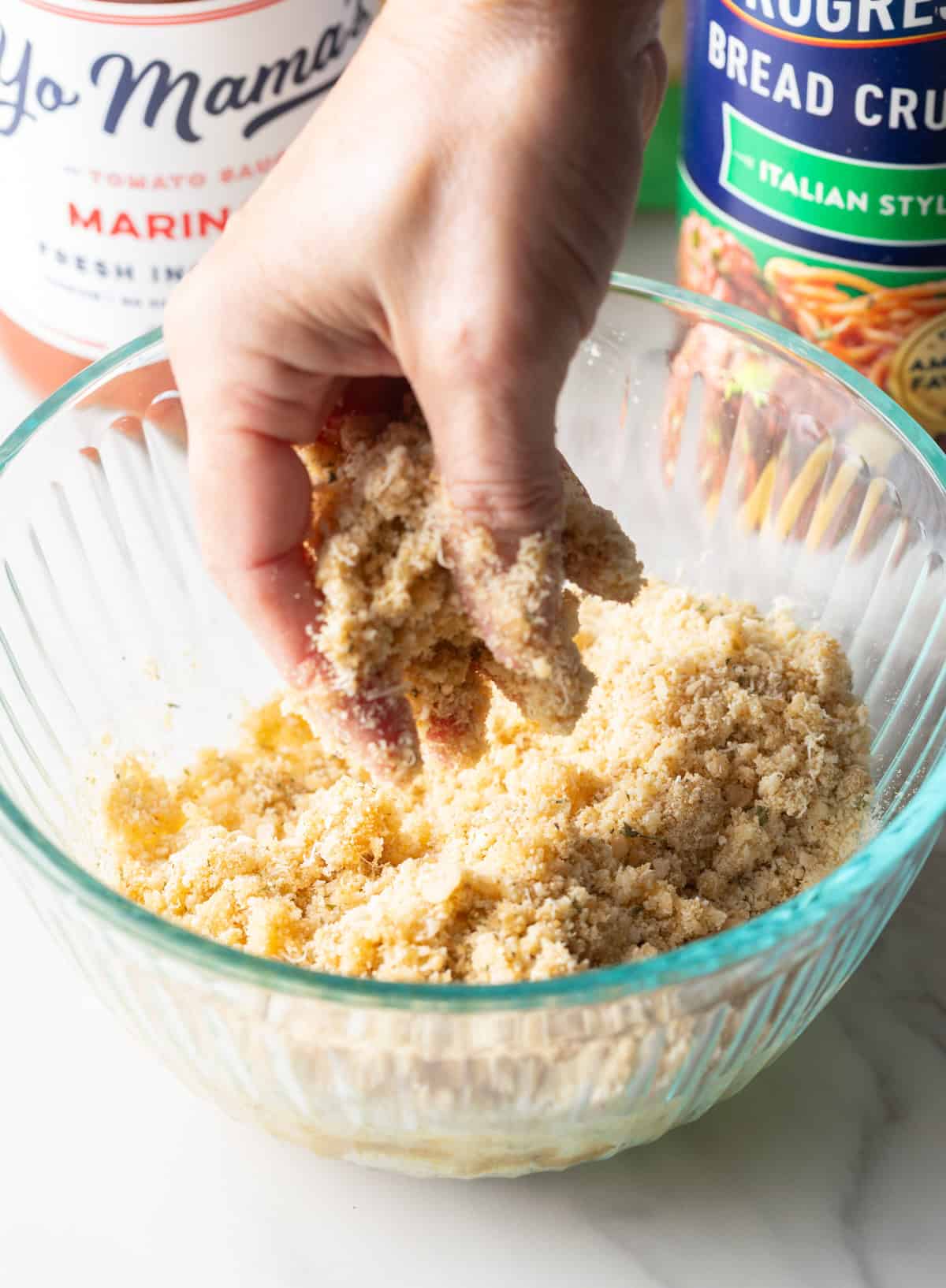 A parmesan and breadcrumb mixture is being mixed by a hand in a glass bowl.