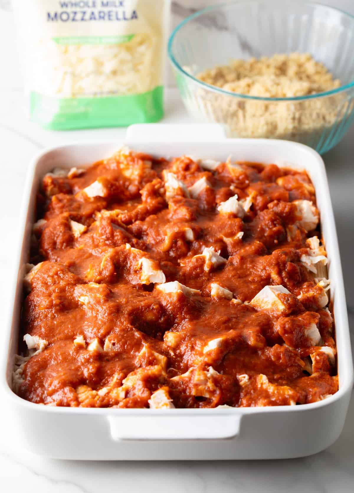 A white baking dish with pasta, chicken, and marinara sauce being prepared for chicken parmesan pasta bake.