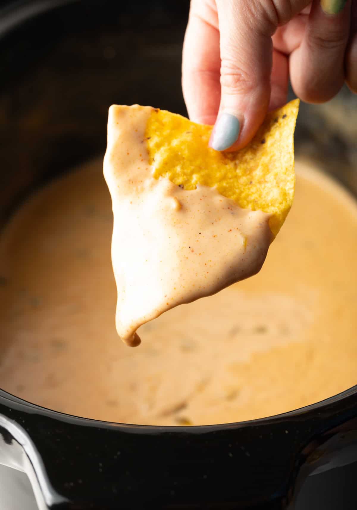 A hand holding a tortilla chip that's been dipped in the crock pot queso recipe hovered over the crock pot.