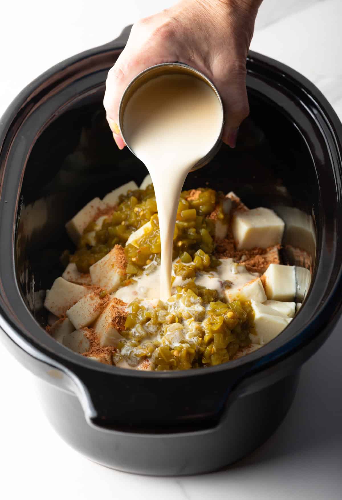 Crock pot queso ingredients assembled in a black crock pot with evaporated milk being poured over it.